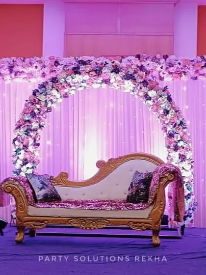 An elegant stage setup for a reception or engagement. The floral arch in shades of purple and white, combined with soft backlighting and a classic chaise lounge, creates a romantic and sophisticated focal point.