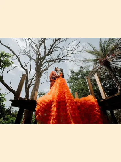 A dramatic, low-angle shot of the couple in orange, the bride's gown cascading down, framed by trees and sky.