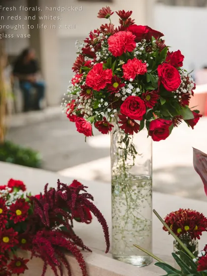 A classic centerpiece of fresh red carnations and roses with baby's breath, beautifully arranged in a tall, clear glass vase.