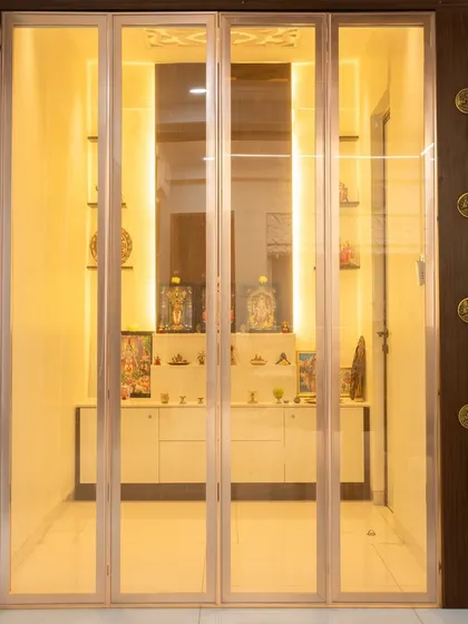 A serene pooja room with calming, gold-tinted glass doors that fold away. The interior is well-lit, creating a peaceful and divine atmosphere.