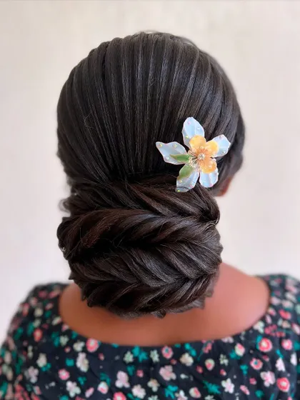A very neat and intricate fishtail braided bun. The look is completed with a single, delicate floral accessory, showcasing minimalist elegance.