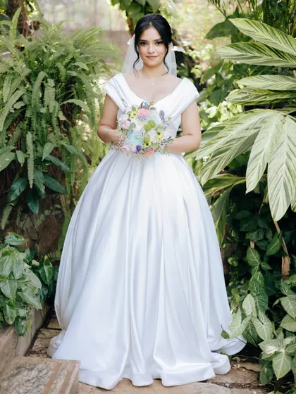 A full-length portrait of the bride in her classic satin ball gown, holding a colorful bouquet in a lush garden.