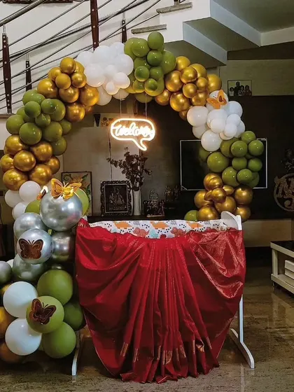 A beautiful and simple cradle decoration for a baby welcoming ceremony at home. The circular balloon arch in green, gold, and white, along with a 'Welcome' neon sign, creates a warm and inviting space.