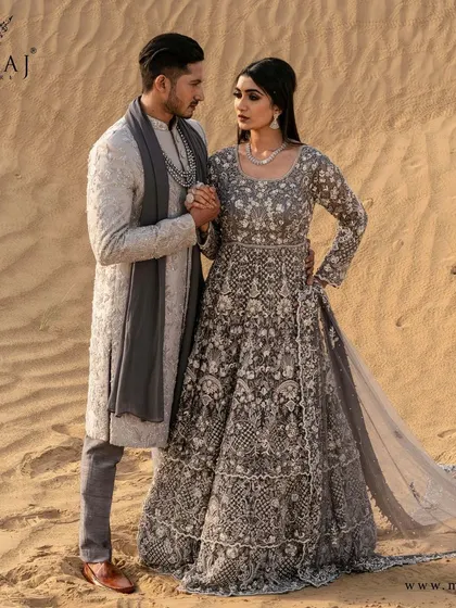 A romantic moment in the desert, showcasing a coordinated Indo-Western look. The groom's grey sherwani with its delicate embroidery holds its own beside the bride's magnificent tiered gown.