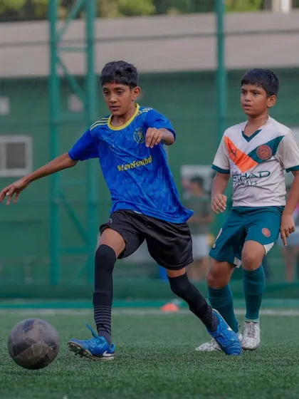A player in a blue jersey drives forward with the ball, showing composure and control under pressure from an opponent. This is a perfect example of where talent meets tenacity.