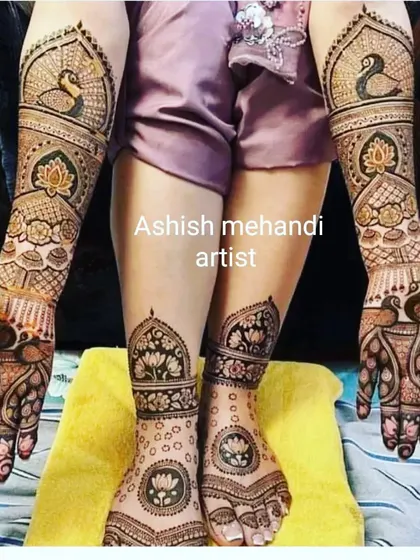 A classic bridal pose showing full coverage mehandi on both hands and feet, featuring lotus and peacock motifs.