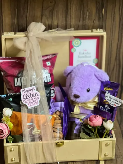 A charming Rakhi hamper in a trunk box, featuring a cute purple teddy bear, chocolates, and snacks, perfect for a beloved sister.
