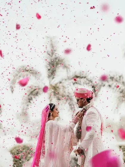 An artistic shot of the couple during their Varmaala, framed by a gentle shower of pink petals against a backdrop of palm trees.