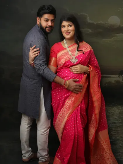 This couple's portrait is set against a painterly backdrop of a moonlit sky. The mom-to-be's bright pink saree and traditional jewelry create a stunning visual against the dark, moody setting.