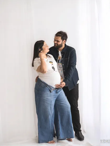 A happy, interactive shot of an expecting couple in casual denim and white tees. This photo captures the genuine smiles and excitement shared between them in a relaxed studio setting.