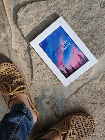 A framed watercolor painting of the Aurora lights. The blend of pink and blue in the sky above the pine tree silhouettes creates a beautiful and dreamy travel-inspired artwork.