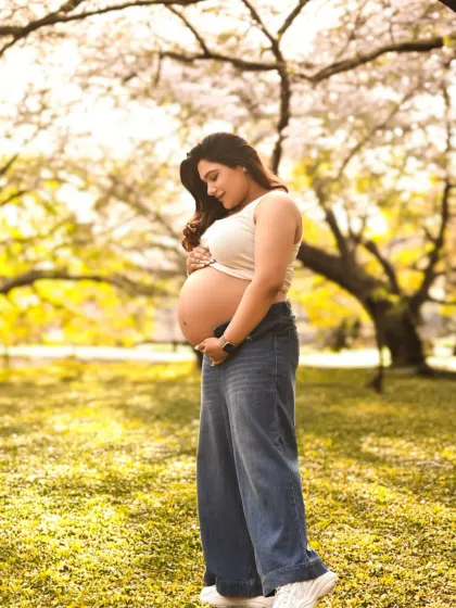 A casual and beautiful outdoor maternity portrait. The expecting mother cradles her bare bump against a backdrop of lush, sunlit trees.
