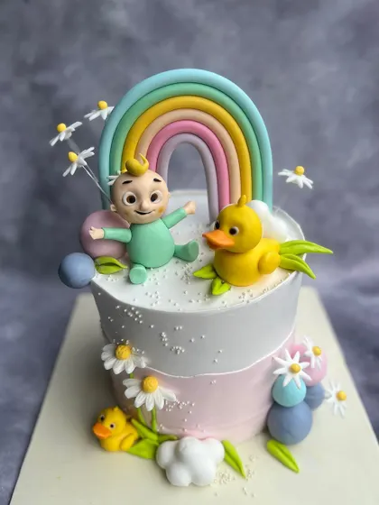 A whimsical Cocomelon cake featuring JJ and his ducky friend under a pastel rainbow. The hand-painted daisies and fluffy fondant clouds add a touch of sweetness to this adorable design.