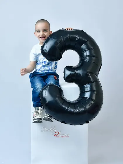 A happy three year old boy celebrating his birthday in the studio. He is sitting on a white block next to a large black balloon in the shape of the number three.