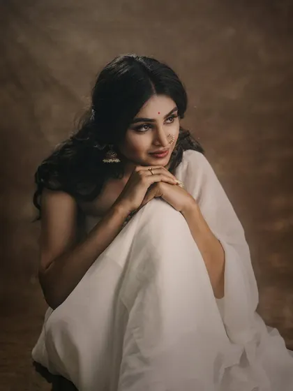 A full-body seated portrait in a white saree against a warm, brown backdrop. The model's thoughtful gaze and the simple styling evoke a sense of classic Indian beauty.