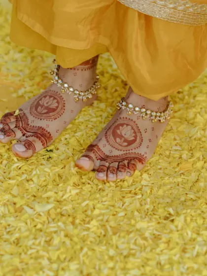 A close-up of the bride's feet, adorned with henna and beautiful anklets, standing on a bed of yellow flower petals.
