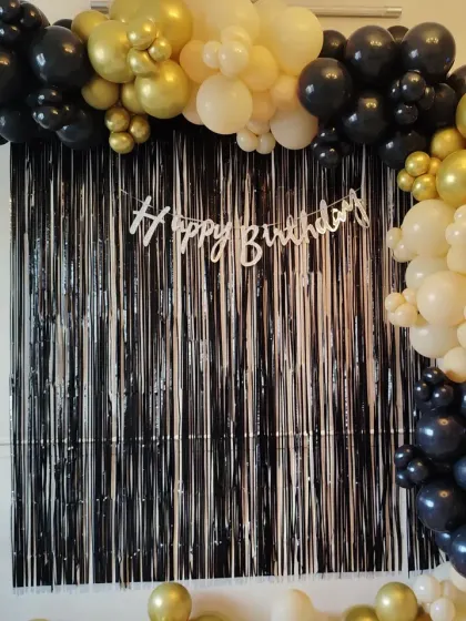 A simple yet stylish home decoration with a black tinsel curtain. The look is completed with a 'Happy Birthday' banner and a black, gold, and white balloon arch.