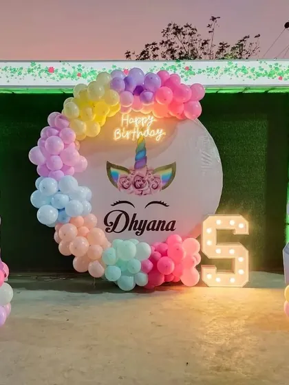 A dreamy unicorn-themed 5th birthday party. This outdoor setup includes a circular backdrop with a unicorn design, a pastel rainbow balloon arch, light-up number '5', and unicorn character cutouts.