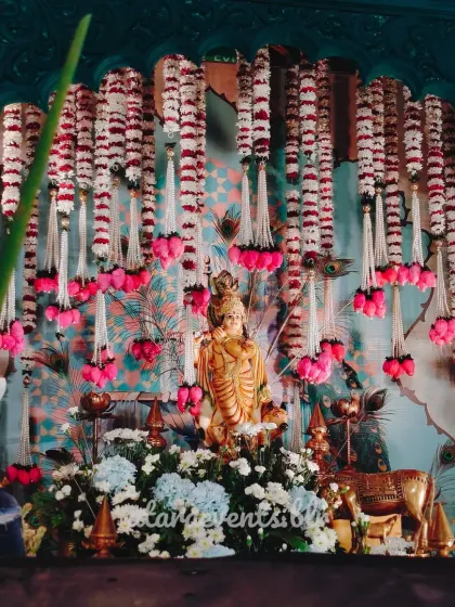 A stunning view of the Krishna-themed stage. The statue is placed centrally, with beautiful garlands of white and pink flowers cascading down, creating a divine and visually impressive focal point.