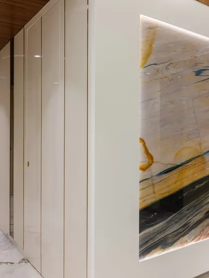 A view of the passage leading to the dining area, where a backlit exotic marble panel creates a dramatic and luxurious entrance. The glossy white doors and wooden ceiling add to the sophisticated feel.