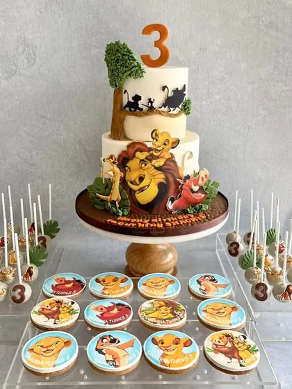 A full dessert table for a Lion King fan! The two-tiered cake features hand-painted scenes from the movie, and it's surrounded by themed cookies and cake pops with all the beloved characters.
