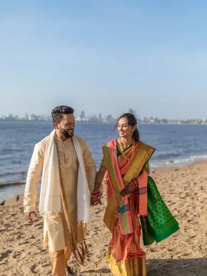 A full-length shot of the couple walking on the beach, capturing the sea, the city skyline, and their happy moment.