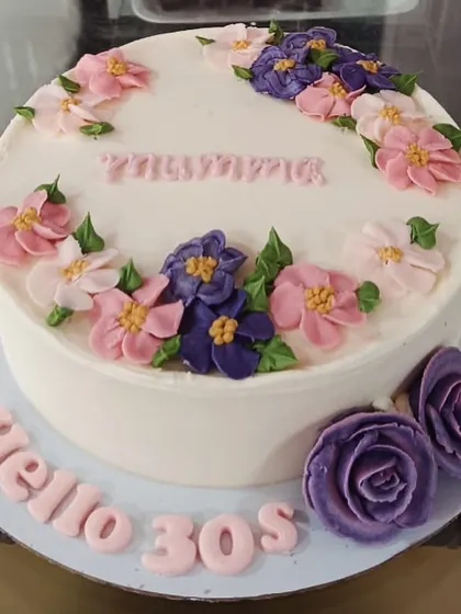 A beautiful floral cake to welcome a new decade, with "Hello 30s" written on the board. The cake is decorated with a crescent of intricately piped buttercream flowers in shades of pink and purple.