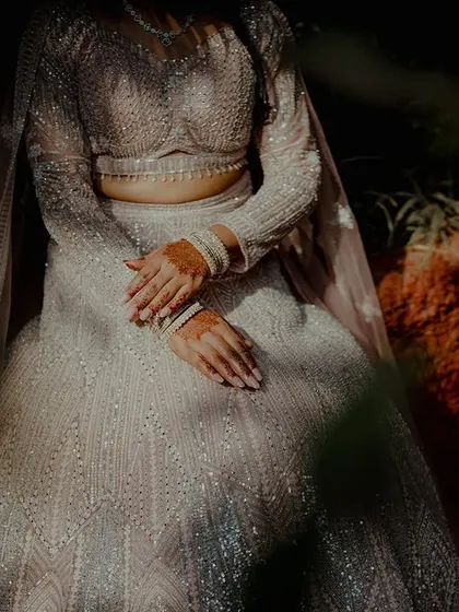 An artistic, moody shot focusing on the hands and shimmering fabric of a bride's lehenga. This detail-oriented photo highlights texture and light.