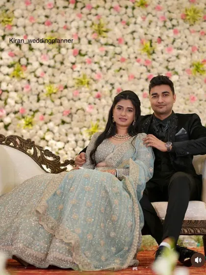 A lovely couple seated on their reception stage, in front of a wall densely packed with white and light pink flowers for a soft, romantic look.