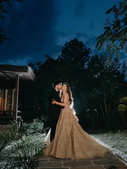 A wide shot capturing the couple in a magical, fairytale-like setting. The twilight sky and the warm lights of the cottage create an enchanting backdrop for their romantic kiss.
