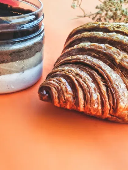 A classic pairing of a flaky, golden-brown croissant and a layered chocolate dessert in a jar. The bright, warm background makes the textures pop, creating a clean and modern look for a cafe menu.