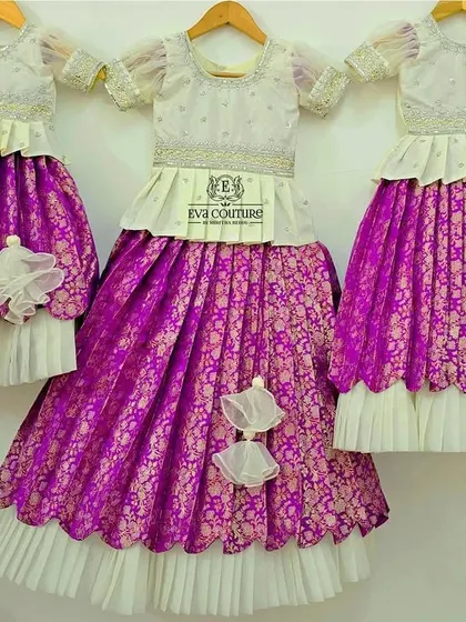 A lovely set of three matching Banarasi silk lehengas, perfect for three sisters. The outfits feature a rich purple brocade skirt and an elegant white peplum top, creating a uniform and graceful look.