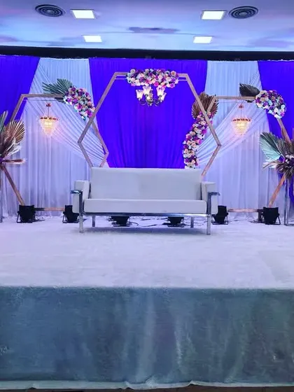 A modern and symmetrical reception stage design featuring two large hexagonal frames. The backdrop combines royal blue and white drapes, with a comfortable white sofa for the couple, creating a contemporary and stylish look.