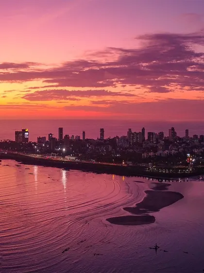 The Mumbai coastline and Marine Drive bathed in the soft, purple and pink hues of a tranquil sunset. This drone shot captures the serene meeting of the city and the Arabian Sea, with the tide creating patterns on the shore.
