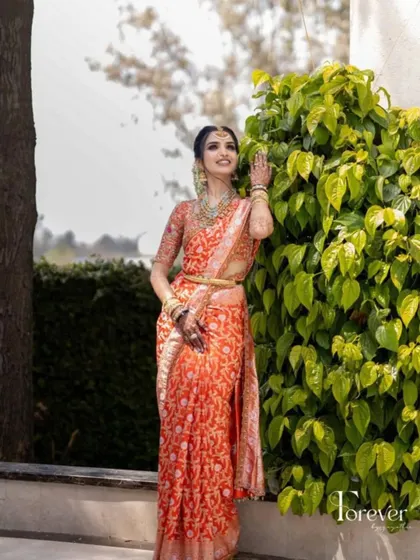The bride posing by lush green leaves, the vibrant orange of her saree creating a beautiful contrast. Her makeup is warm and radiant.