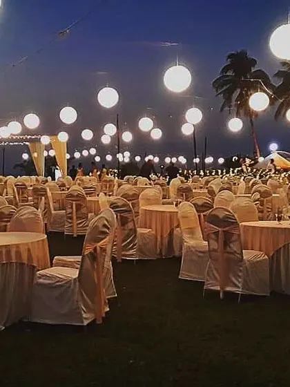 An evening view of a lawn reception setup. The large, glowing paper lanterns hanging over the guest tables create a beautiful and magical atmosphere under the night sky.