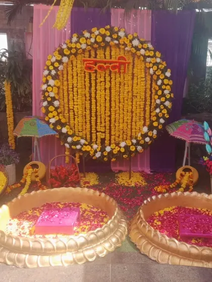A vibrant Haldi setup with two lotus-shaped urlis filled with flower petals. The backdrop is a large ring of marigolds, decorated with small umbrellas and colorful drapes.