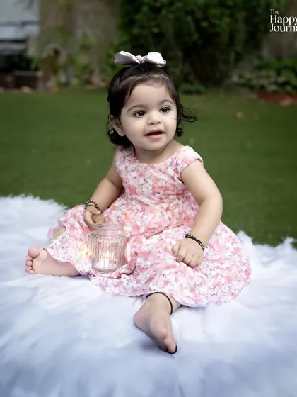 A toddler sits on a fluffy white blanket outdoors, holding a candle jar. The soft light and her gentle expression create a peaceful scene.