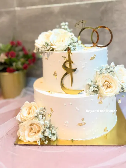 This elegant two-tier Nikkah cake features a clean white finish with scattered gold leaf. It's personalized with a custom Arabic initial topper and adorned with beautiful off-white roses and baby's breath.