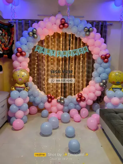 A classic ring backdrop for a baby shower, featuring a mix of pink and blue balloons with metallic accents. A "Baby Shower" banner is hung in the center against a curtain of warm fairy lights.