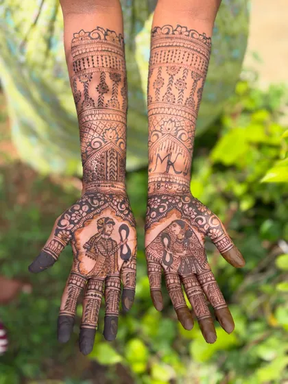 A clear view of a full bridal mehendi featuring bride and groom portraits. The design is balanced with traditional patterns like jhumkas and architectural elements on the forearms.