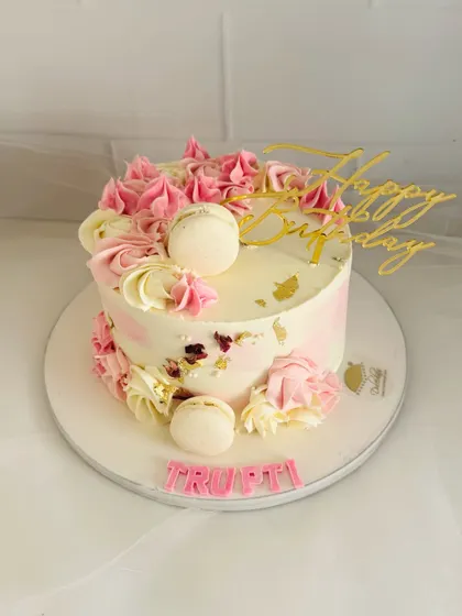 An elegant birthday cake with pink and white buttercream swirls, delicate macarons, and a touch of gold leaf.