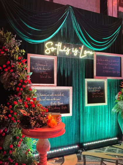 "So This Is Love." A romantic and moody photo-op corner, with a neon sign, framed love quotes, and a backdrop of dark green velvet.