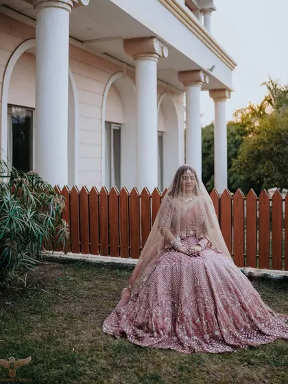 A wide-angle portrait that sets a royal scene. The bride is seated in the gardens of a large, pillared building, her voluminous lehenga spread out like a queen's gown.