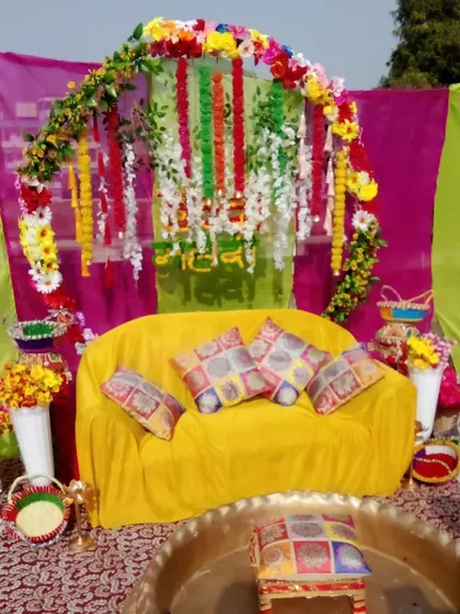 An outdoor Mehendi setup with a simple floral arch over a yellow sofa. The decor uses a mix of colorful drapes and props like birdcages for a rustic feel.