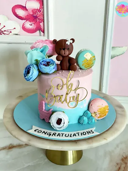 A sweet "Oh Baby" cake for a baby shower, featuring a teddy bear, macarons, and beautiful blue and pink flowers.