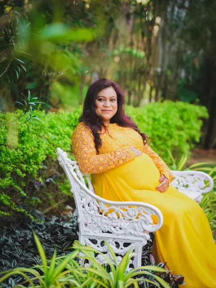 A peaceful portrait of a mother-to-be in a lovely yellow gown, seated on a white wrought-iron bench in a verdant garden.