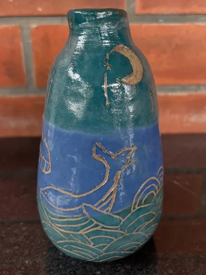 This vase, featuring a whale tail and a crescent moon, was hand-built and carved by a 12-year-old student. The sgraffito technique reveals the lighter clay body underneath the blue glaze.