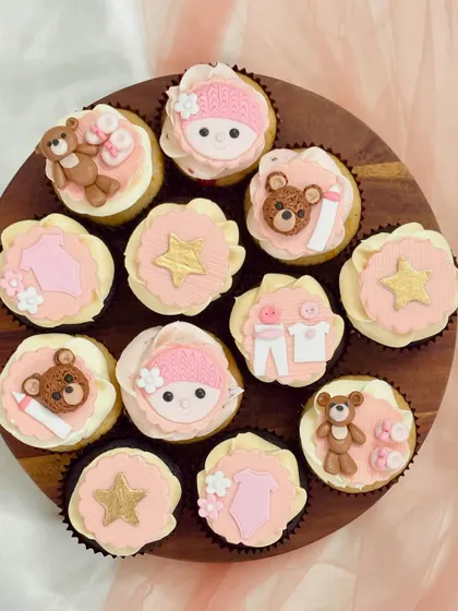 An assortment of pink-themed baby shower cupcakes on a wooden board. The designs include fondant teddy bears, baby clothes, gold stars, and baby faces with knitted hats, demonstrating a cohesive and detailed theme for celebrating a baby girl.