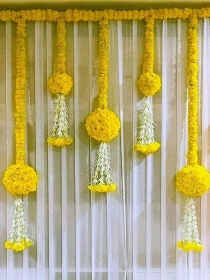 Simple details can make a big impact. For this Haldi background, we created an elegant look using hanging floral tassels made from marigold balls and white jasmine flowers against a sheer white curtain.